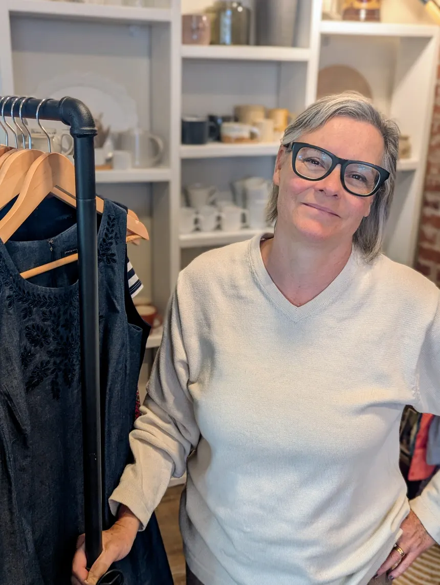 Clare Rivard, owner of The Village Collective consignment boutique in Canning, Nova Scotia, standing in her shop beside a rack of consigned clothing.